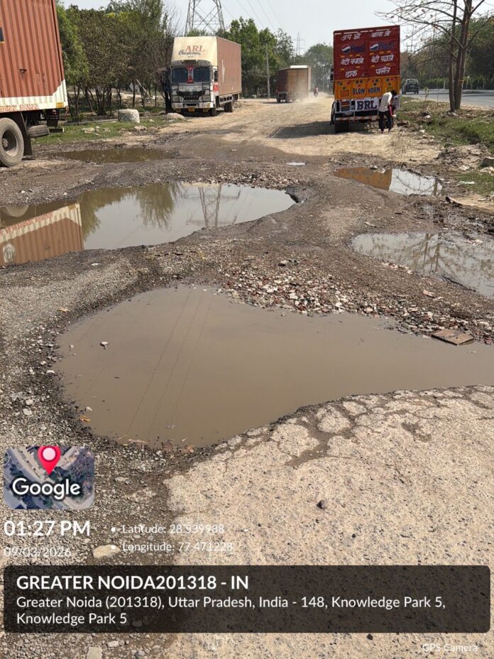Waterlogged roads and massive potholes in Knowledge Park 5 expose severe infrastructure neglect and ongoing drainage issues in Greater Noida.