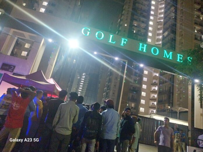 Angry residents gather at the society gate late at night, protesting against the alleged security scam as police arrive to manage the situation.